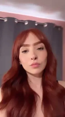 A woman with red hair talking on a video chat from her room