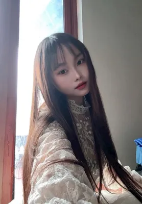 A woman with long dark hair in a lace top taking a selfie by a window
